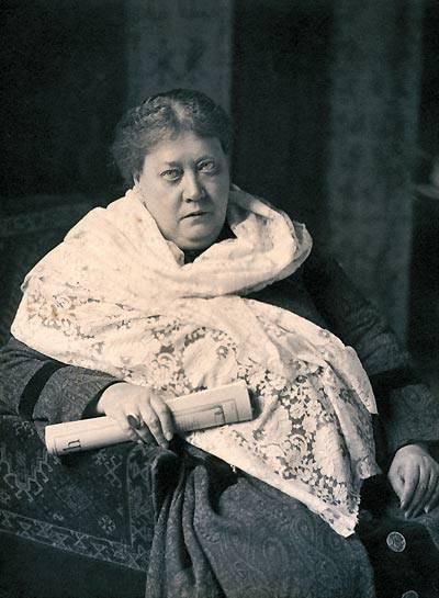 Retrato fotográfico de Helena Blavatsky sentada, envolta num xale claro de renda, segurando um rolo de papel sobre o colo e olhando diretamente para a câmera.